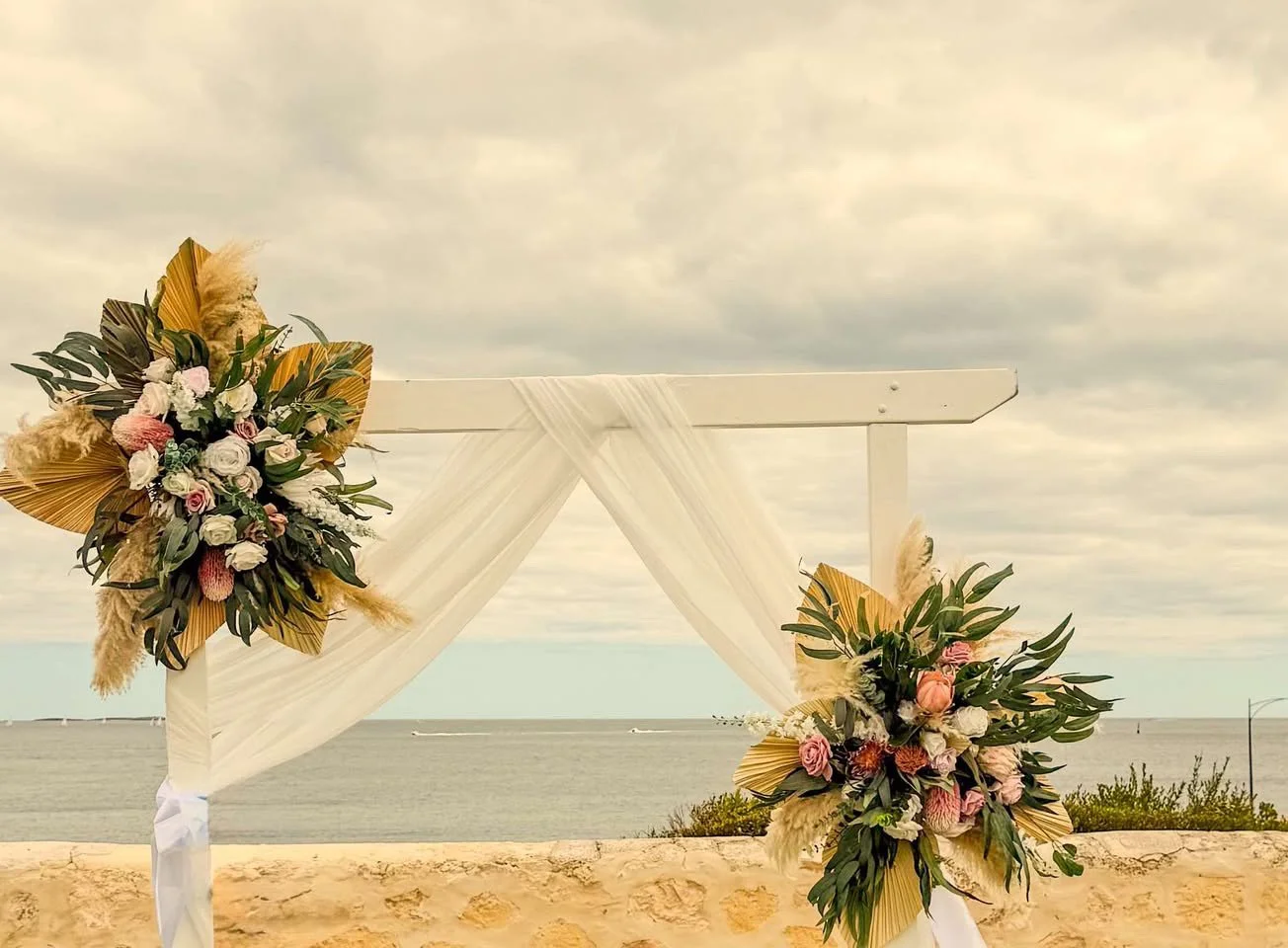 A wedding altar structure decorated with floral arrangements of pink and white flowers, green leaves, and dried pampas grass, set against the beach and ocean with a cloudy sky.