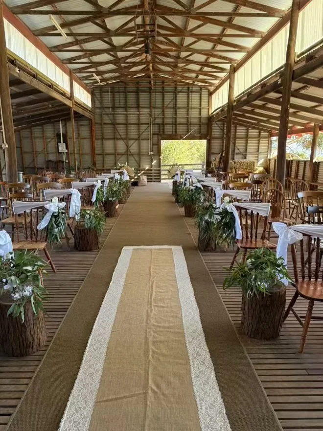 Indoor rustic wedding venue with wooden chairs decorated with white ribbons and greenery, a beige aisle runner with lace borders, and natural light coming from open sides of a barn-style structure.