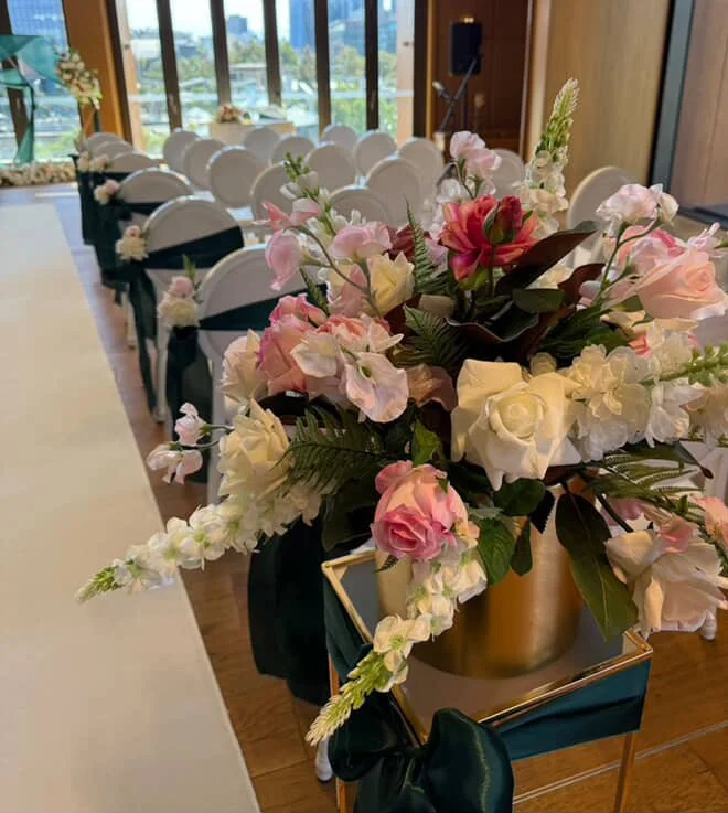 A large floral arrangement with pink and white roses, pink and white snapdragons, and green foliage in a golden vase at a wedding or event setup.