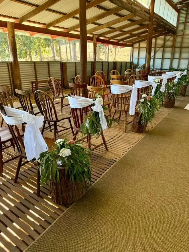 Wedding or event seating area with wooden chairs decorated with white fabric sashes and floral arrangements in wooden pots along the aisle, under a rustic, open-air shelter.