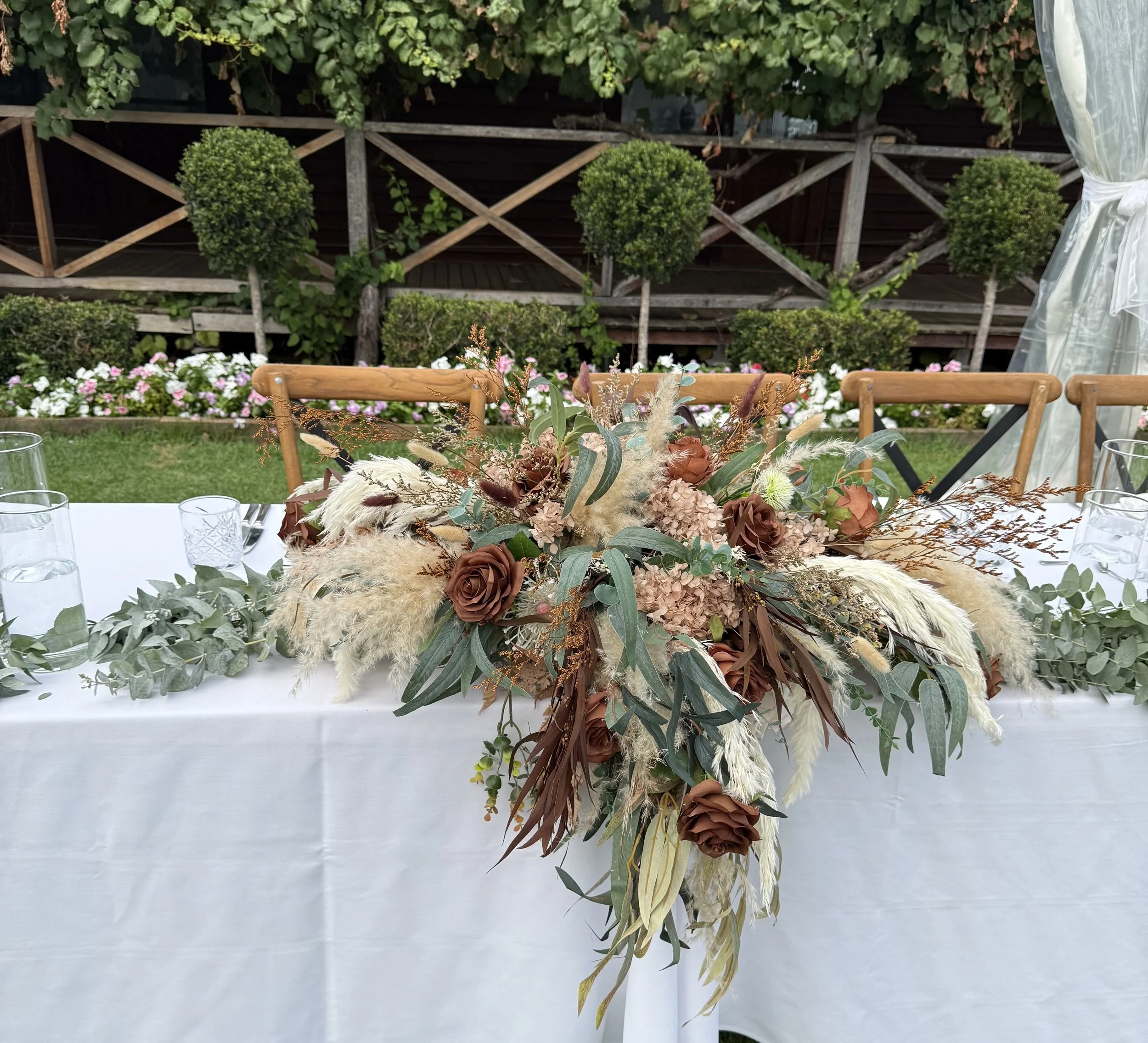 A floral centerpiece with various flowers and greenery on a white table, with a garden and wooden fence in the background.