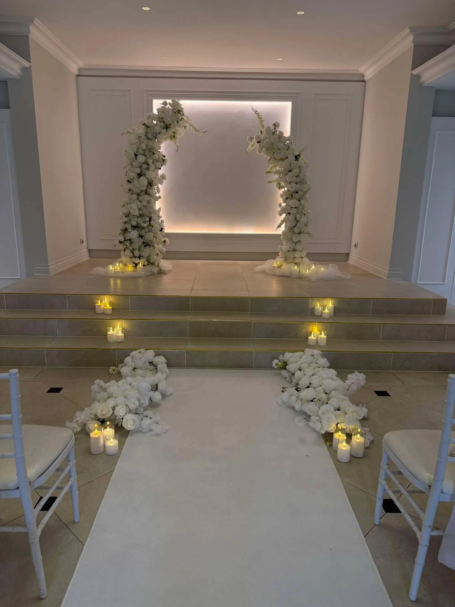 A wedding ceremony setup with a white floral arch, candles, white chairs, and a white aisle runner in a decorated indoor venue.