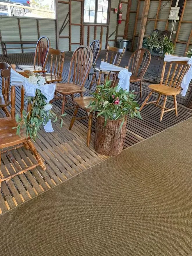 Indoor rustic dining area with wood chairs decorated with white fabric and greenery, a log planter with flowers, and a wooden slat floor.