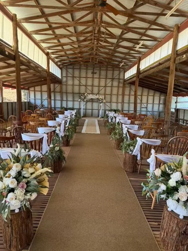 Wedding ceremony setup inside a rustic barn with wooden chairs, floral arrangements, and an arch at the altar.