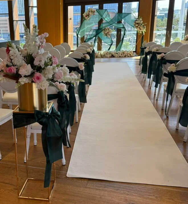 Wedding ceremony setup with floral arrangements along aisle, decorated chairs with black ribbons, modern art installation at the altar, and large windows in the background.