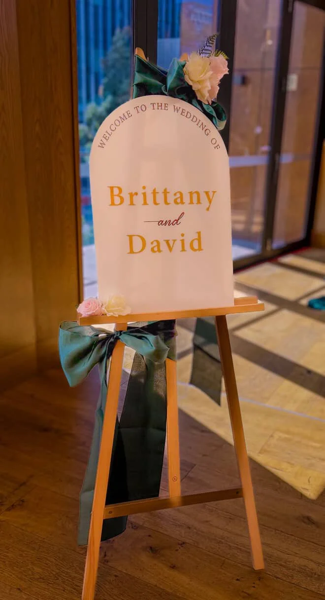 Wedding sign on an easel reading, 'Welcome to the wedding of Brittany and David,' decorated with flowers and a ribbon, situated near a doorway with wooden flooring and glass doors in the background.