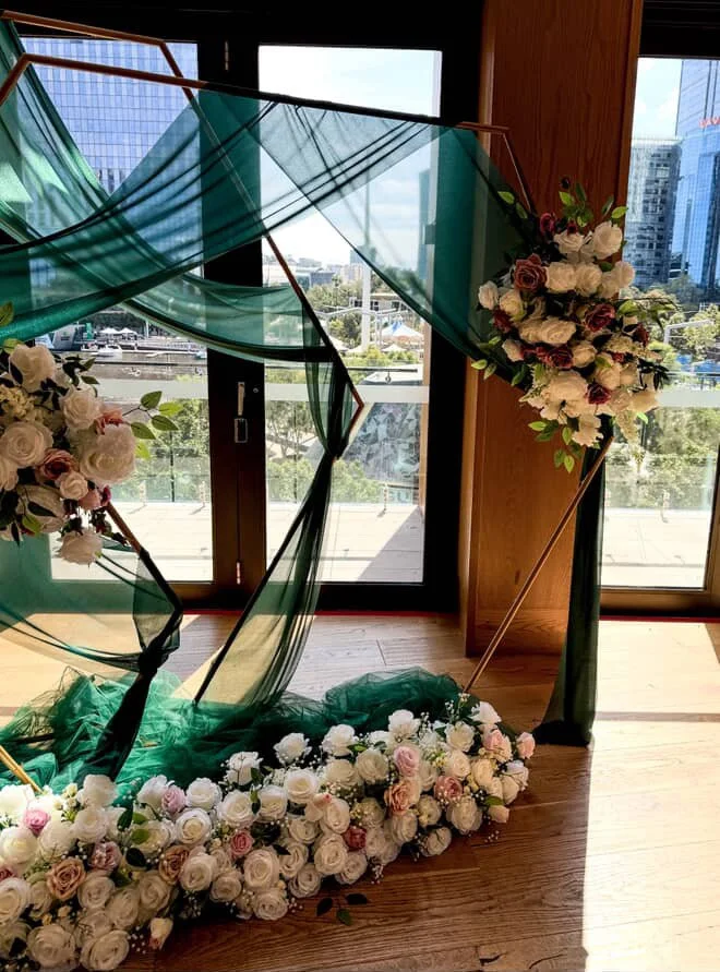 A floral wedding or event backdrop featuring white, pink, and purple roses, green foliage, green tulle fabric, and wooden geometric structures on a wooden floor by large glass doors with a city view.