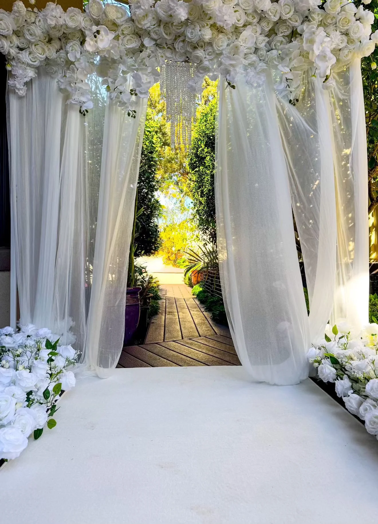 White floral wedding arch with curtains and hanging crystals, overlooking a garden pathway with trees and sunlight.