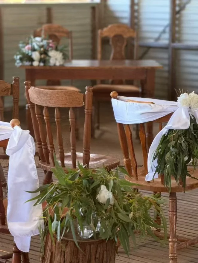 Rustic wedding or event setup with wooden chairs decorated with white fabric and flowers, and a floral arrangement on a log in the foreground.
