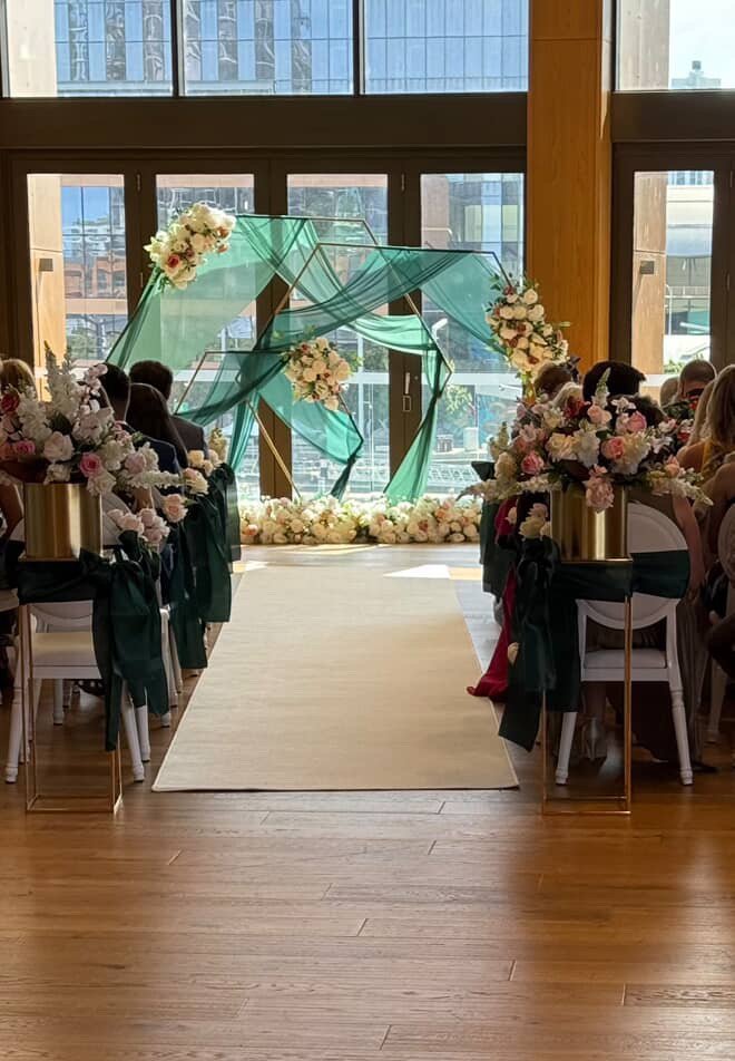 Indoor wedding ceremony setup with an aisle leading to a flower arch and decorative drapery in front of large windows.