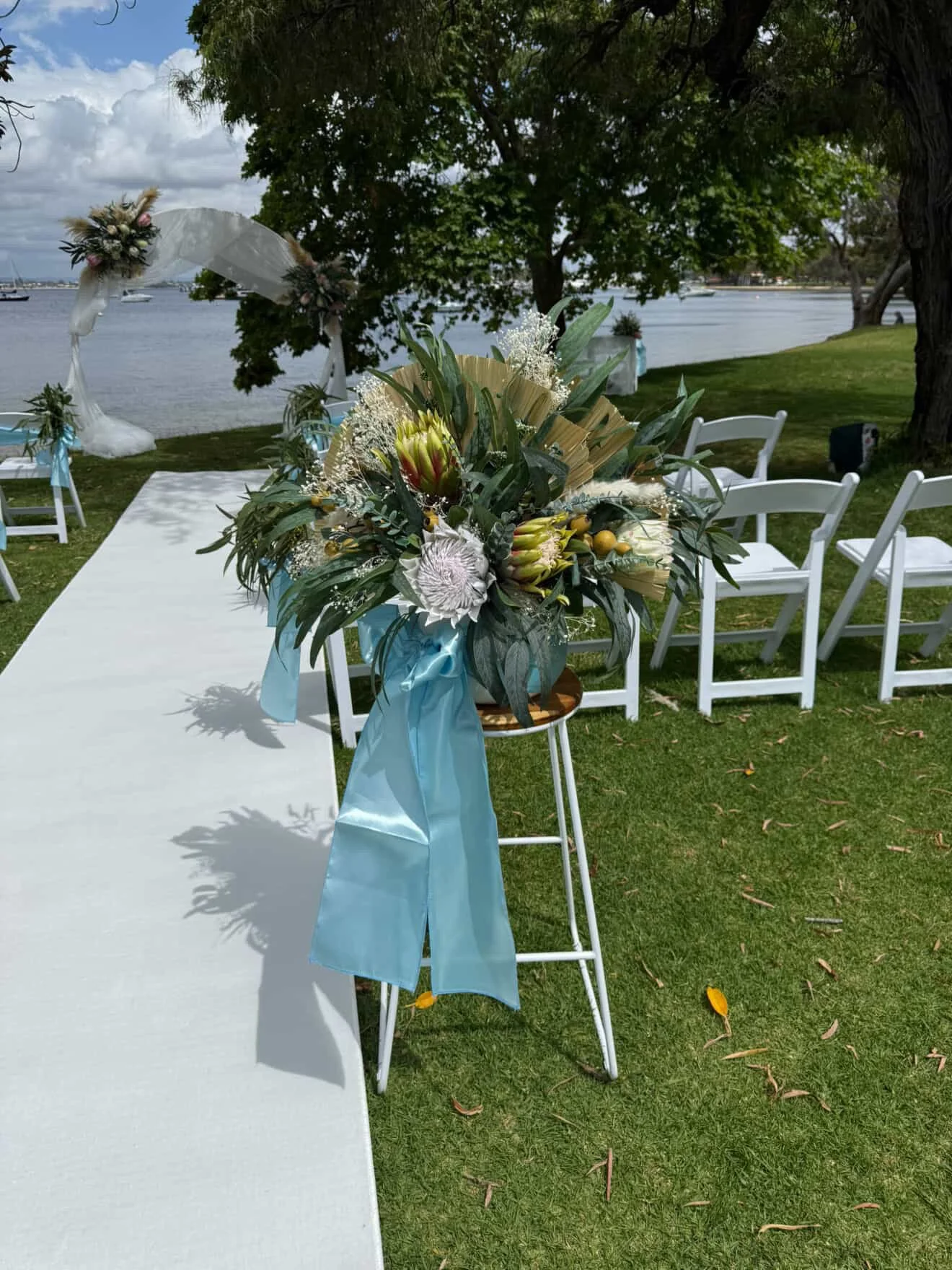 A decorated outdoor wedding ceremony setup by a river, featuring white chairs, a floral arrangement with greenery and flowers, and a wedding arch decorated with flowers and fabric, surrounded by trees and a cloudy sky.