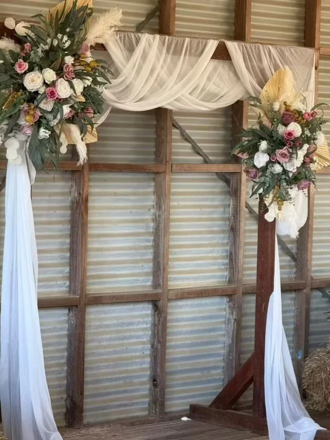 Wooden rustic frame decorated with floral arrangements and white fabric, set against a corrugated metal wall.