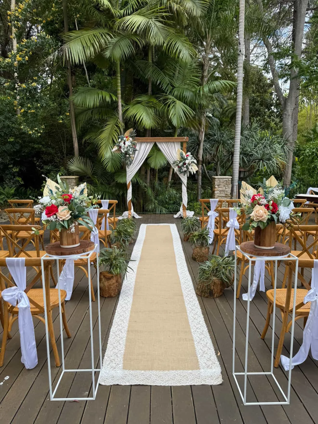 Outdoor wedding ceremony setup with wooden chairs decorated with white fabric ribbons, floral arrangements, and a beige aisle runner leading to a wooden arch draped with white fabric and floral arrangements, surrounded by lush green tropical foliage.