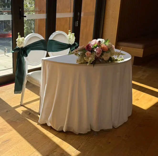 A round table with a white tablecloth and a floral centerpiece, positioned near glass doors with sunlight streaming in, with two white chairs decorated with dark teal ribbons tied into bows.
