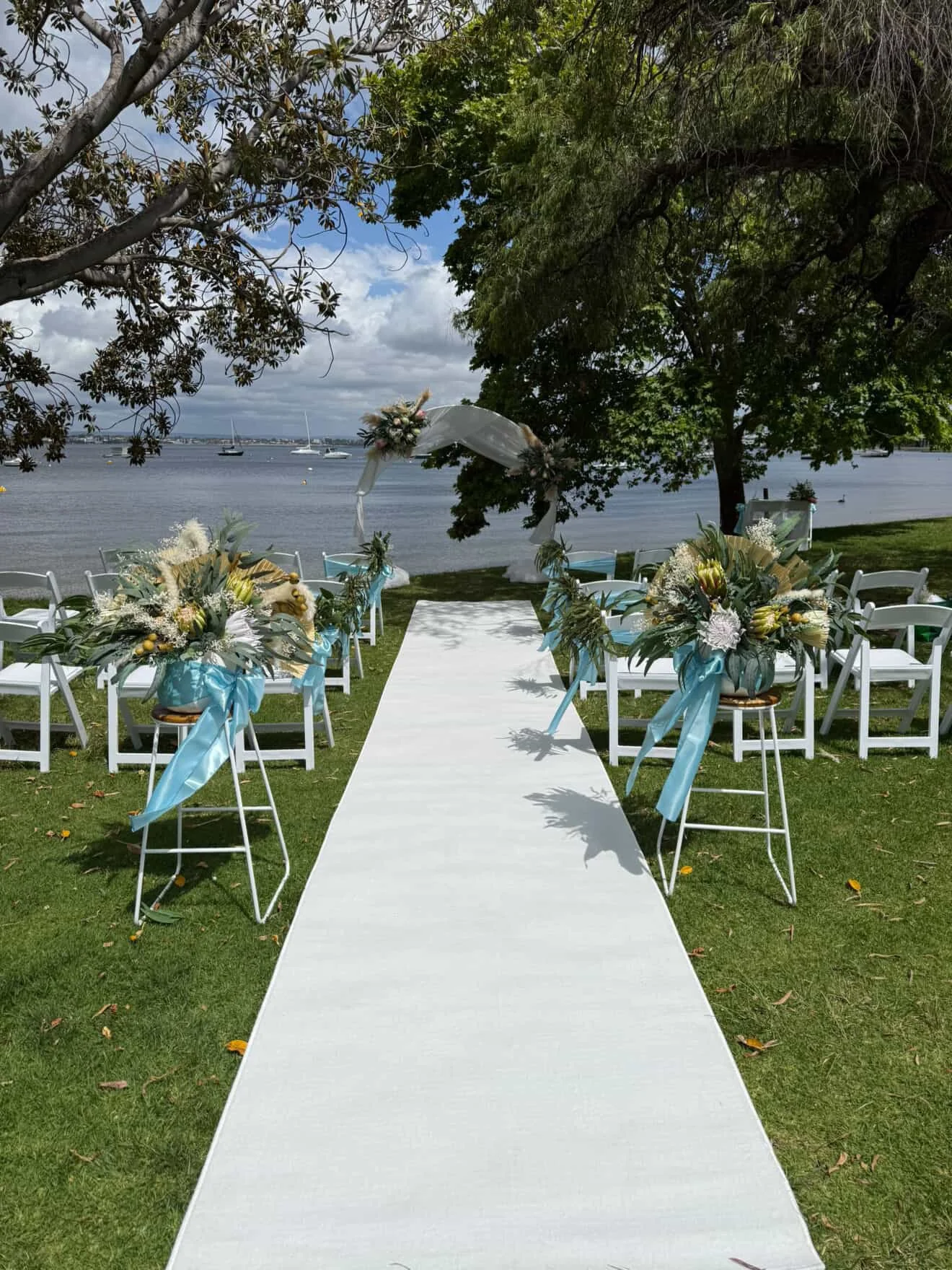 A beachside wedding setup with a white aisle runner, white chairs decorated with blue ribbons and floral arrangements, and a floral arch overlooking the water with sailboats, trees, and cloudy sky in the background.