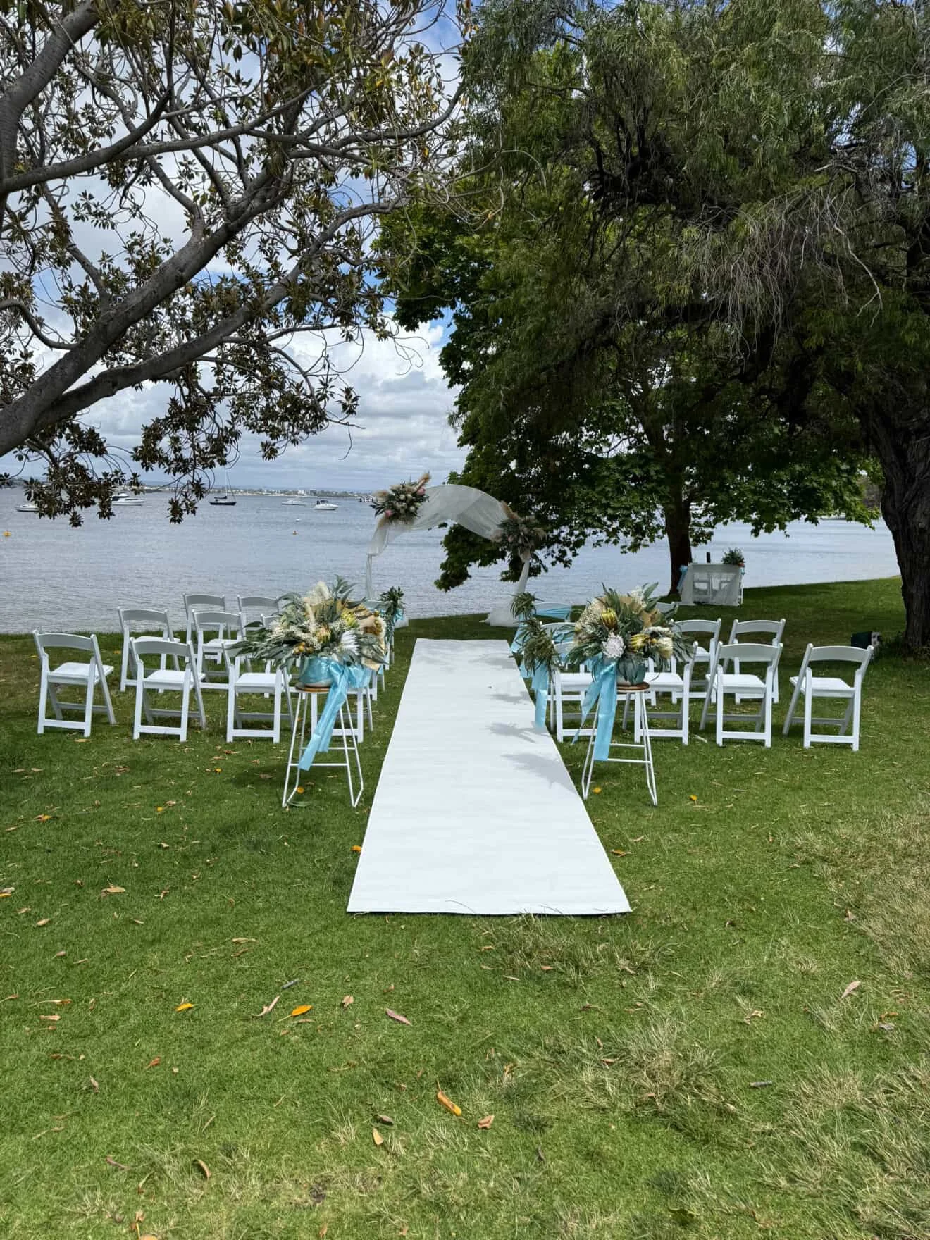 Outdoor wedding setup on a grassy area by a body of water, with white chairs arranged along a white aisle runner, decorative flowers, trees, and sailboats in the background under a partly cloudy sky.