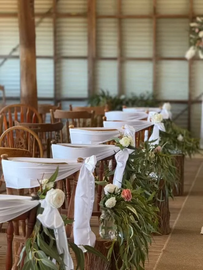 Wedding chairs decorated with white fabric and flowers in jars, arranged along a wooden platform in a rustic setting.