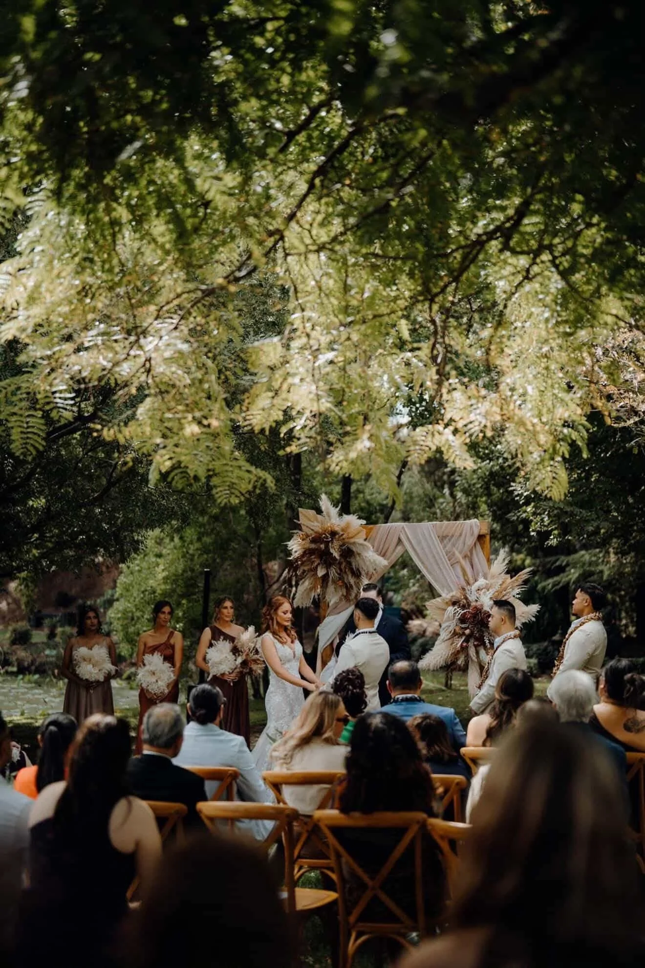 A wedding ceremony taking place outdoors beneath a canopy of green trees, with the bride and groom holding hands and exchanging vows, surrounded by bridesmaids and groomsmen, with guests seated in front.
