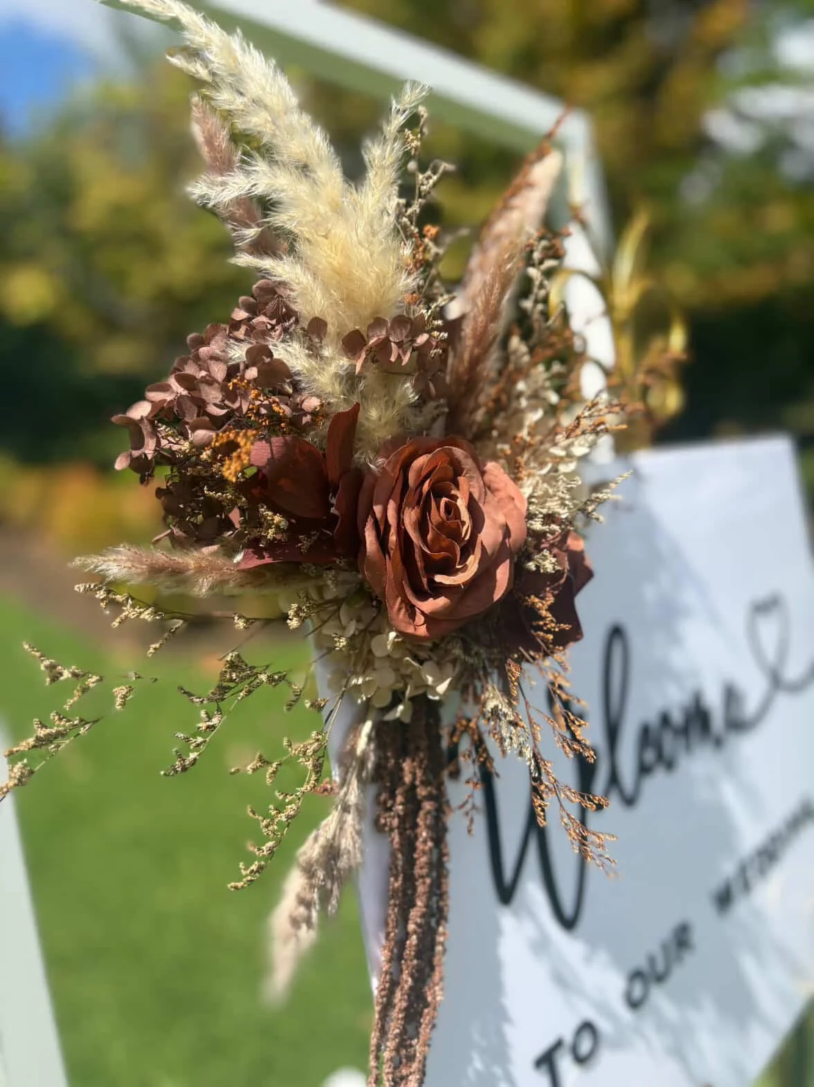 An autumn-themed floral arrangement with a large brown rose, dried grasses, and leaves, attached to a white sign with black text at an outdoor event.