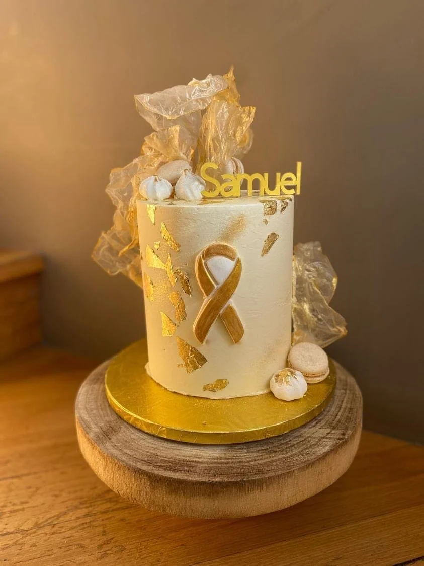 A small, cylindrical cake decorated with gold leaf accents, white icing, and a gold ribbon, topped with a gold name tag that says 'Samuel' and topped with delicate gold and white decorations. The cake sits on a gold cake board on a wooden stand.