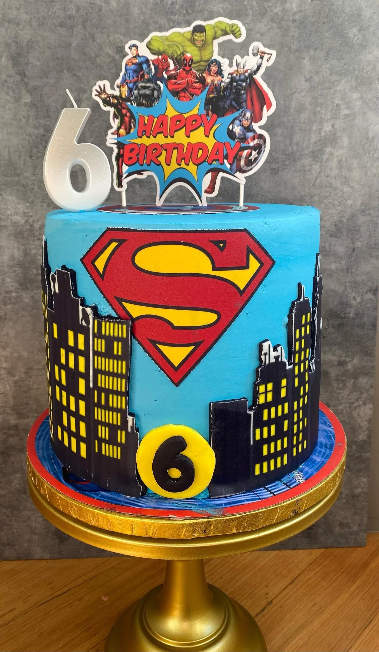 A birthday cake decorated with superhero and city skyline themes, featuring a Superman logo, a large yellow and black number 6, and a "Happy Birthday" sign with Marvel superheroes.