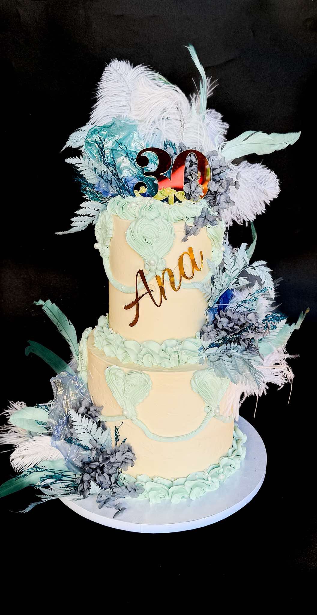 A multi-tiered white cake decorated with feathers, leaves, and flowers, with a "30" birthday topper and the name "Anah" written on it.