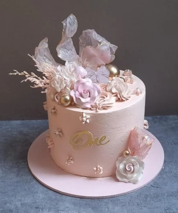 Pink birthday cake decorated with roses, pearls, and gold accents, with the word 'One' written on it.