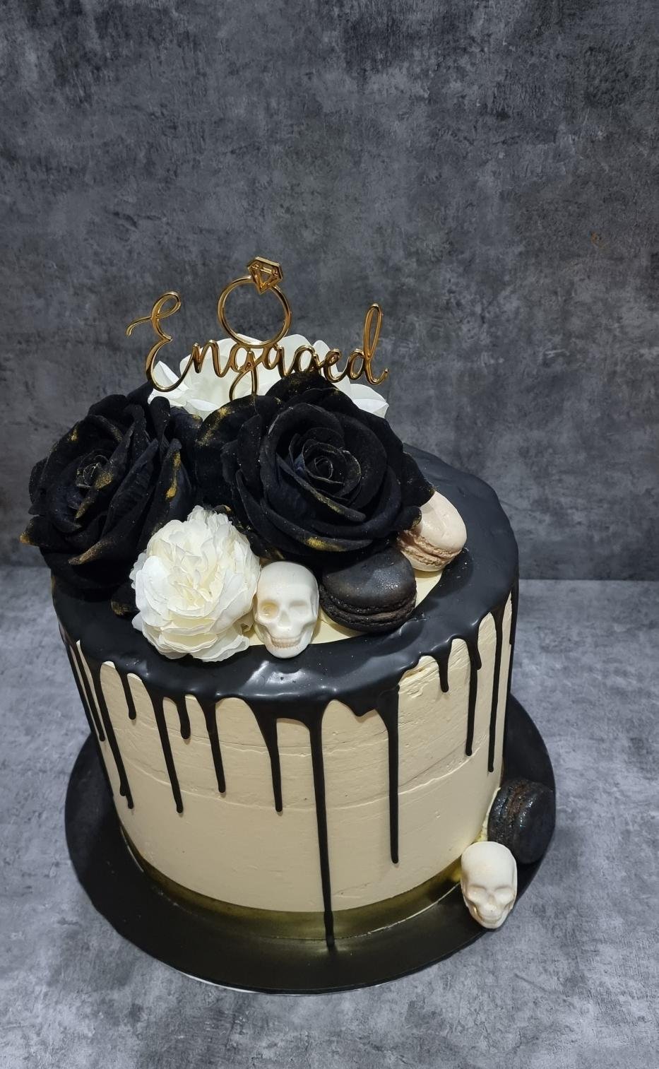 Black and beige birthday cake decorated with black roses, white flowers, skulls, macarons, and a golden "engaged" cake topper featuring a ring and a diamond