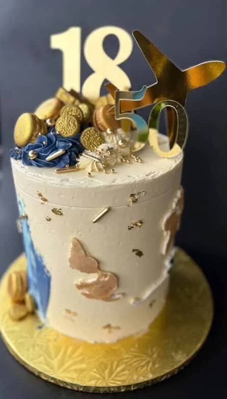 Celebration cake with gold, blue, and silver decorations, topped with a gold dog silhouette, number candles '18' and '50', and various sweets, on a gold cake board.