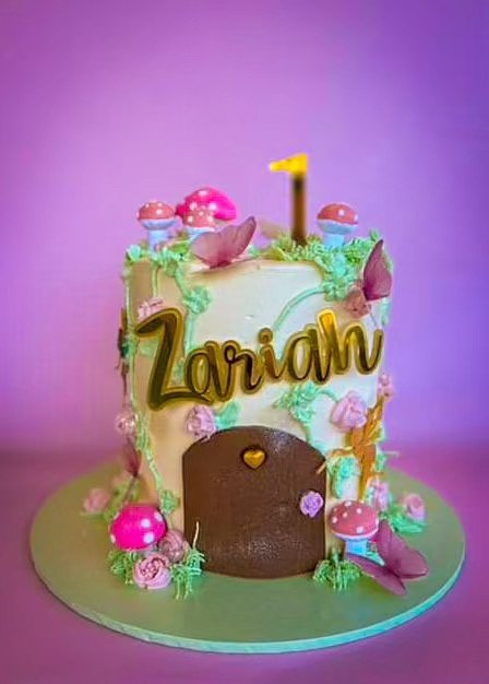 Colorful birthday cake with pink and purple mushrooms, flowers, and green decorations, topped with a yellow number one candle, personalized with the name 'Zariah' on the front.