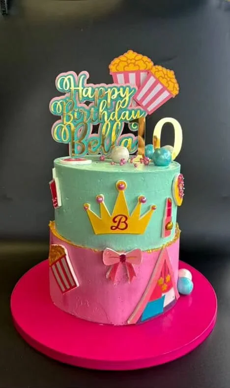 Two-tiered birthday cake decorated with pink and green frosting, a gold crown with the letter 'B', colorful sprinkles, a pink bow, and cartoon popcorn and candy decorations on top. The cake has a 'Happy Birthday Bella' sign and is placed on a pink ca