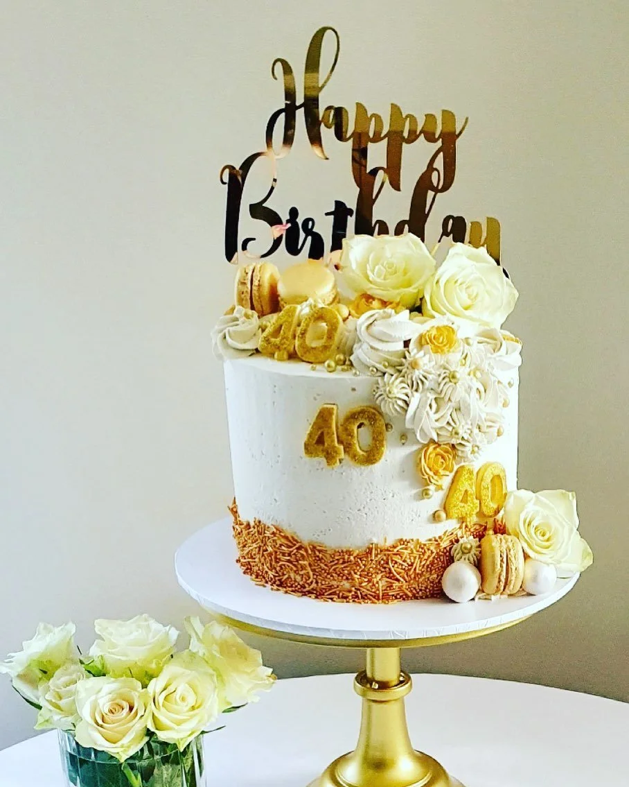 A white birthday cake decorated with white roses, gold and white macarons, and golden number '40' toppers. The cake has a 'Happy Birthday' topper and is displayed on a gold stand. A small bouquet of white roses is in a glass vase next to the cake.