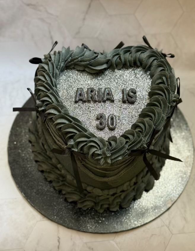 Chocolate heart-shaped cake with silver glitter and black piped icing, decorated with black ribbons, with the message "ARIA IS 30" on top.