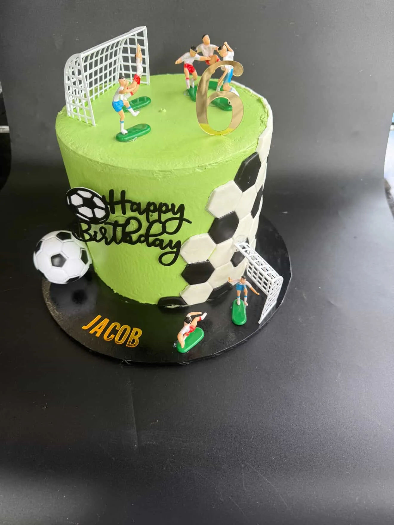 Green birthday cake with soccer theme decorations, including miniature soccer players, a soccer ball, and soccer goal models. The cake has a black base with a yellow name tag reading 'JACOB' and the phrase 'Happy Birthday' with a soccer ball graphic.