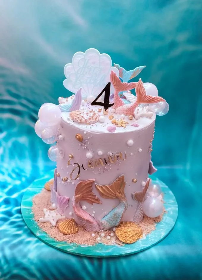A decorated birthday cake with an ocean theme, featuring seashells, mermaid tail decorations, pearls, and pink starfish, on a blue ocean background. The cake has the name 'Jemery' and the number 4 on top.