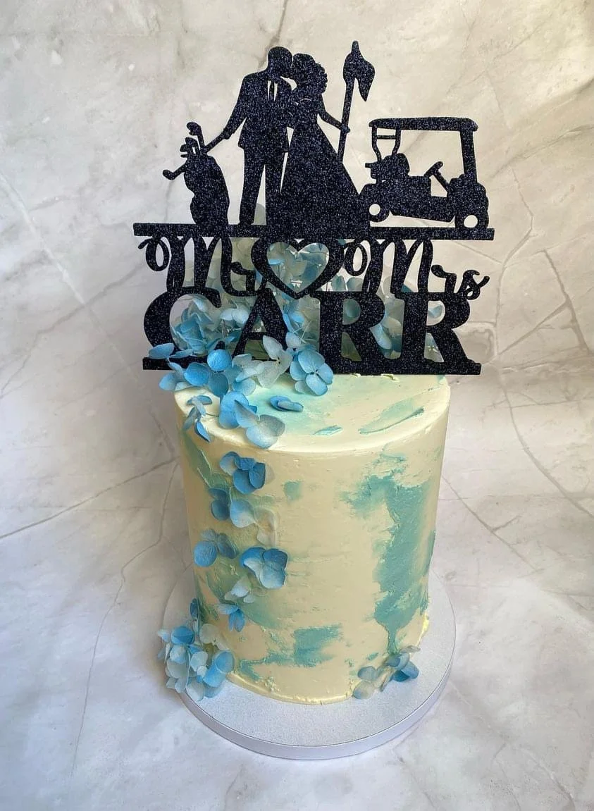 A wedding cake with a black topper that reads  Mr & Mrs, featuring silhouettes of a bride and groom, a golf cart, and golf clubs, with blue and white floral decorations.