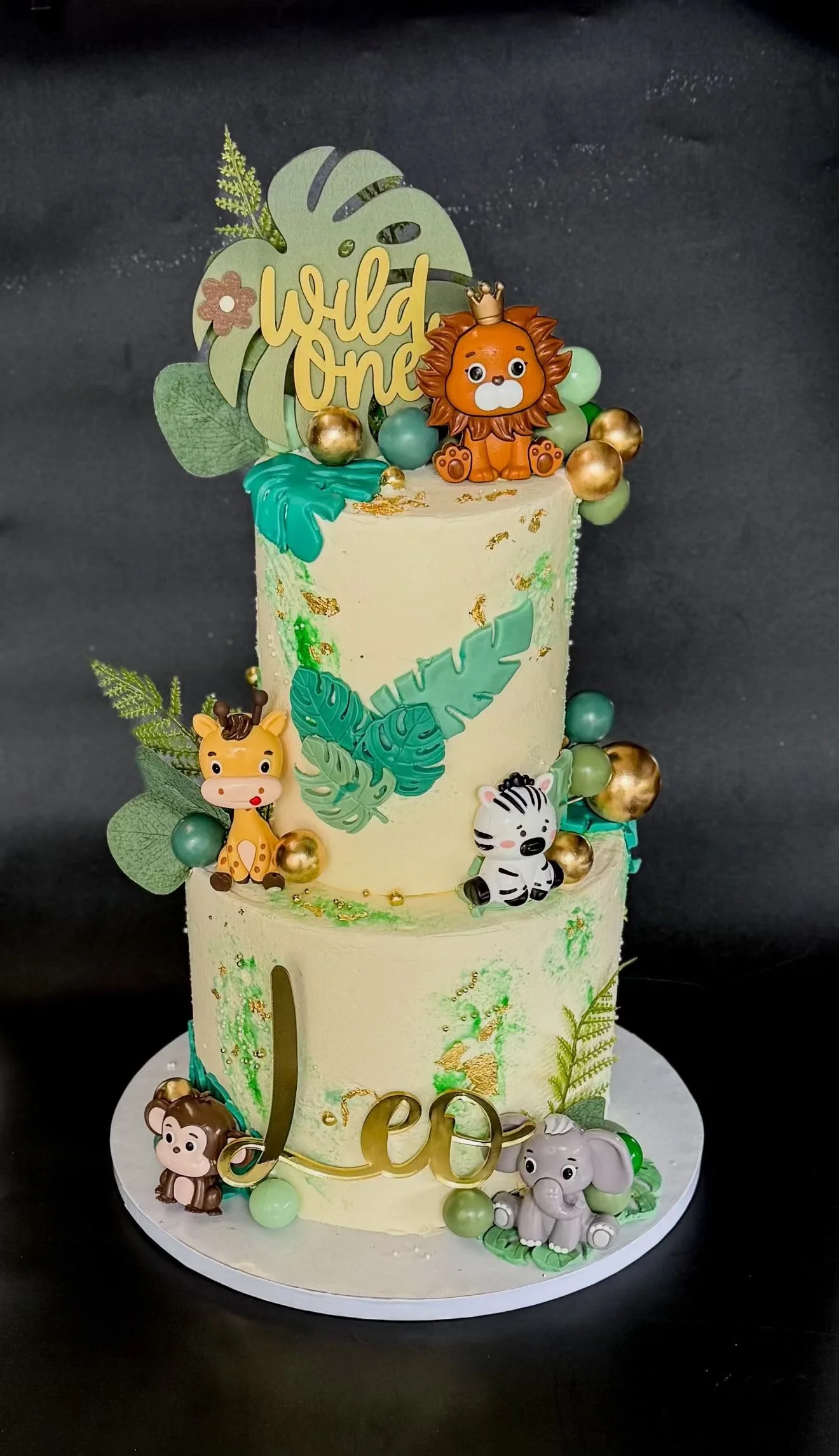 Three-tiered birthday cake decorated with jungle animal and leaf-themed figurines and decorations, including a lion, zebra, giraffe, monkey, and elephant, with a "world One" topper and gold and green embellishments.