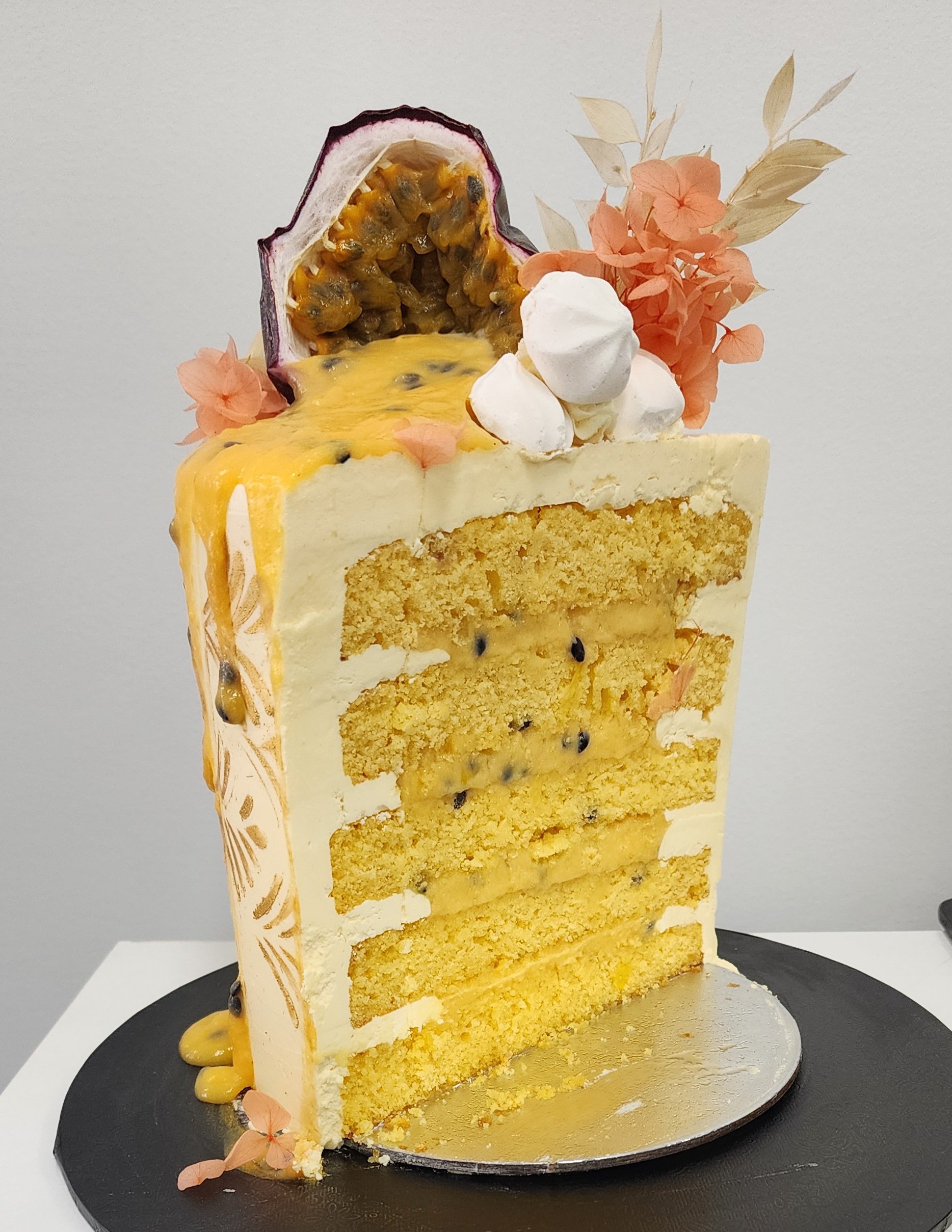 A sliced passion fruit-topped layered cake with white and yellow layers, decorated with whipped cream, dried flowers, and passion fruit pulp on a black cake board.
