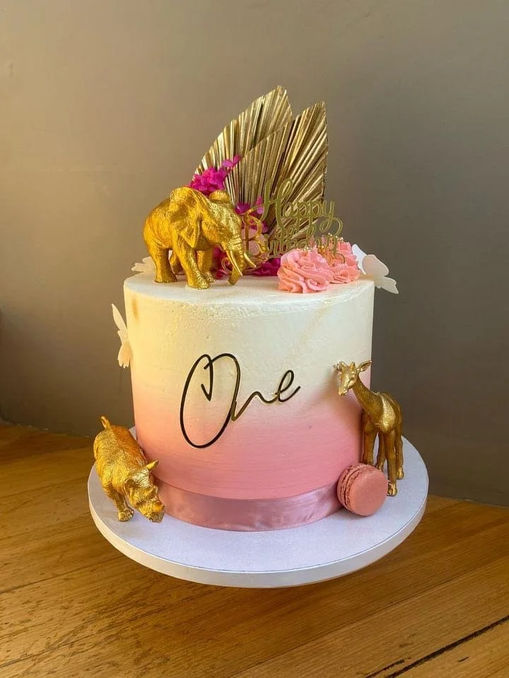 A decorated birthday cake with gold elephant figurines, pink flowers, gold leaves, and a 'Happy Birthday' topper. The cake is white with a pink gradient and the word 'One' written on it, celebrating a first birthday.