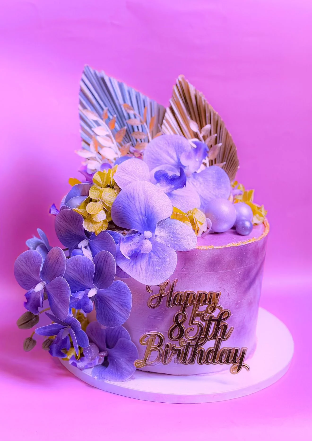 A pink-colored birthday cake decorated with purple and yellow flowers, gold leaves, and pearl-like spheres, with the words 'Happy 85th Birthday' written on the side.