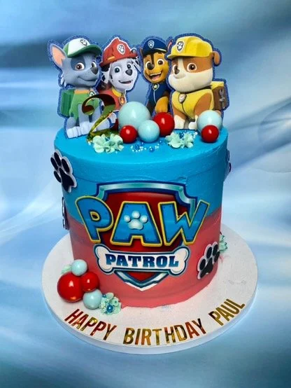 A Paw Patrol themed birthday cake with characters Chase, Marshall, Rubble, and Rocky on top, decorated with blue and red balls and paw print designs, on a white cake board with a birthday message.