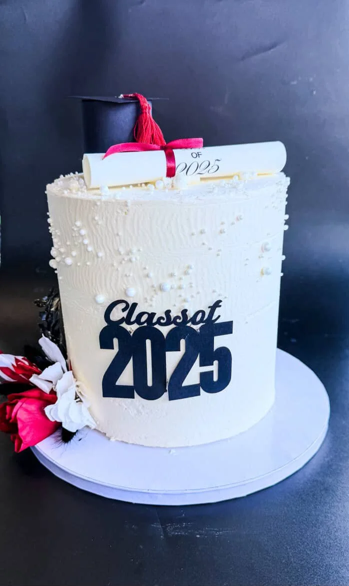 White graduation cake with black "Class of 2025" topper and a small black graduation cap and red tassel on top.