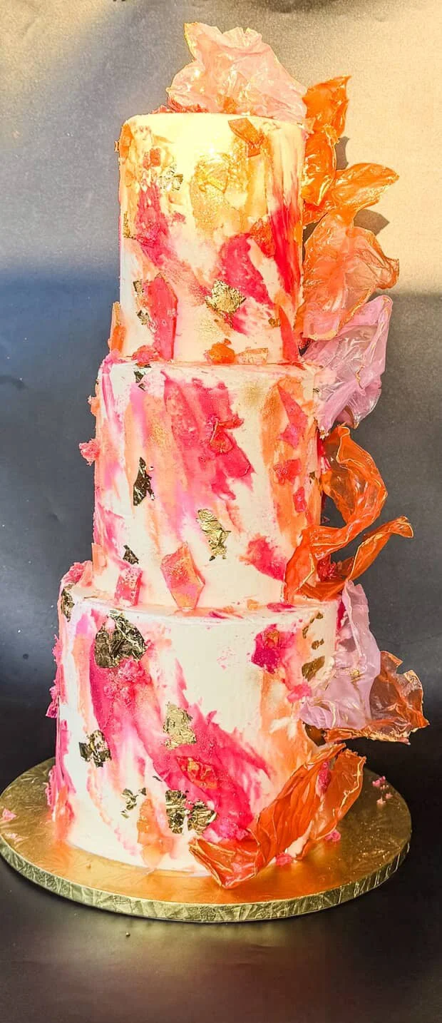 A three-tiered wedding cake with pink, orange, and gold accents, decorated with irregularly shaped edible gold leaf and pink and orange edible flowers.