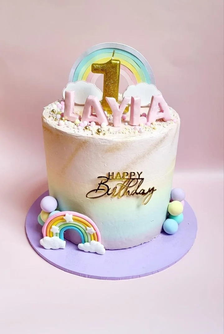 A pastel-colored birthday cake decorated with rainbow, clouds, and balls, with a large gold number one candle, pink letters spelling 'LAYA,' and a gold 'Happy Birthday' topper, set against a pink background.