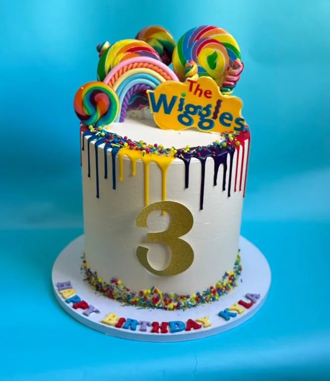 Colorful birthday cake topped with lollipop decorations, rainbow drizzle, and a yellow sign reading 'The Wiggles'. The cake has a gold number 3 in the center, with multicolored sprinkles and a birthday message around the base. The background is blue.