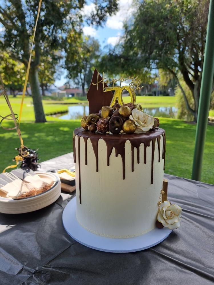 A white birthday cake with chocolate drip decoration on top, adorned with gold-themed decorations, a large number 70 candle, white roses, and a wooden topper that says 'Happy 70'.
