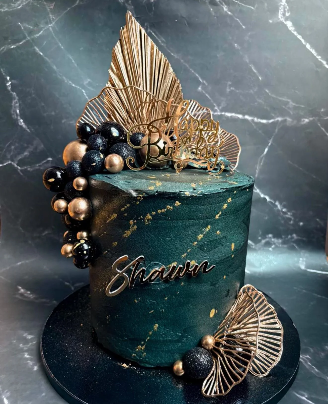 A teal-colored birthday cake decorated with gold accents, black and gold spherical decorations, and large gold leaves. The cake has a "Happy Birthday" topper and a name "Shaun" on the side, set against a dark marble background.