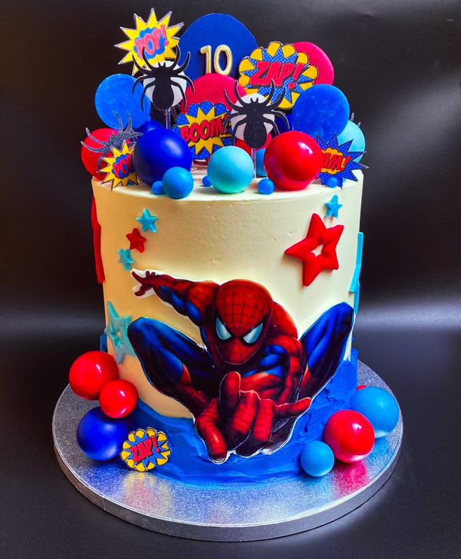 Spider-Man themed birthday cake with colorful comic book style decorations, including stars, speech bubbles with words like "WOW," "ZAP," and "BOOM," and a large spider symbol on top, placed on a silver cake board.