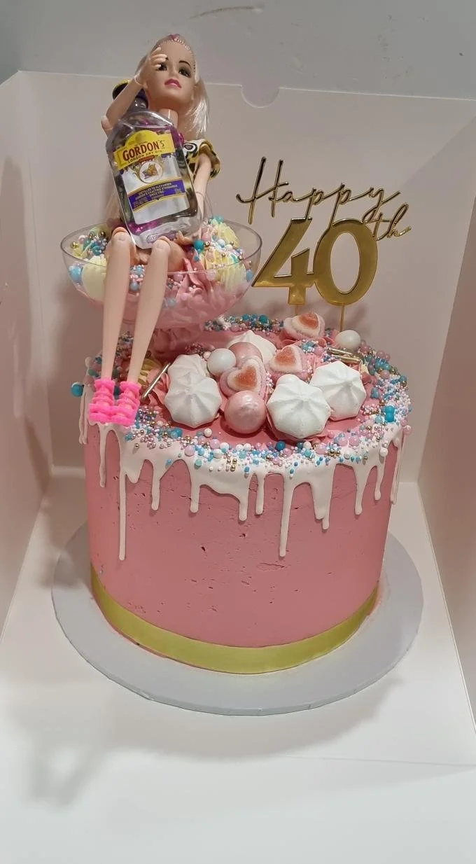 Pink birthday cake with white drip icing, rainbow sprinkles, meringue cookies, candies, and a Barbie doll figure holding a bottle on top, with a gold "Happy 40th" cake topper.