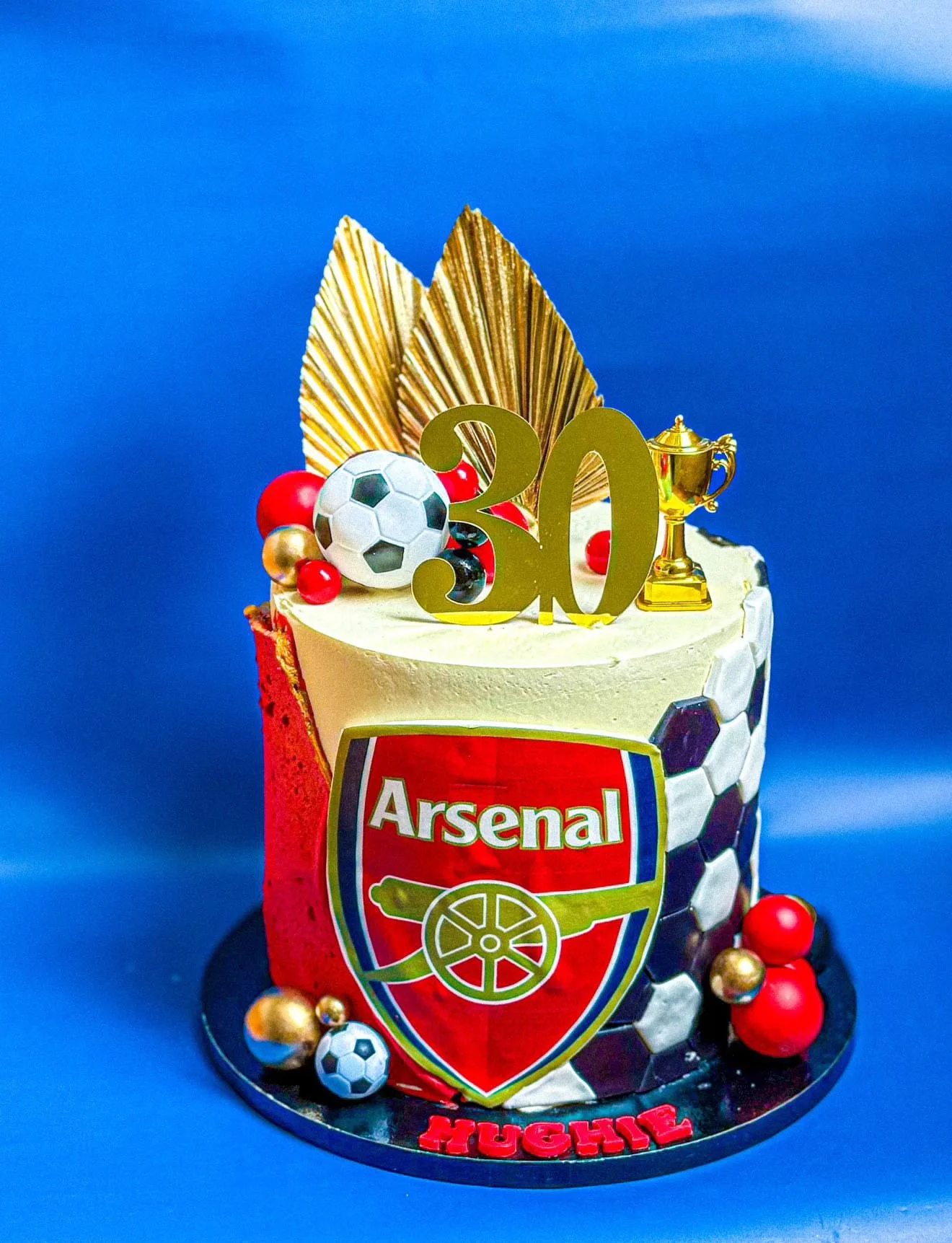 Soccer-themed birthday cake decorated with a large gold '30' number, gold fan-like decorations, a soccer ball, a gold trophy, red and gold spheres, and a 'Hugo' name topper, with a soccer ball pattern on the cake's sides.